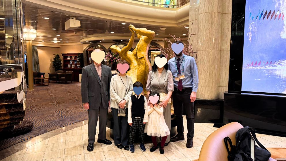 飛鳥Ⅱのアトリウムにある、華やかなゴールドのダンスをするカップル像の前で撮影した、3世代家族6人の記念写真。全員がフォーマルな服装で笑顔。右側の巨大LEDスクリーンには海と船の映像が映っている