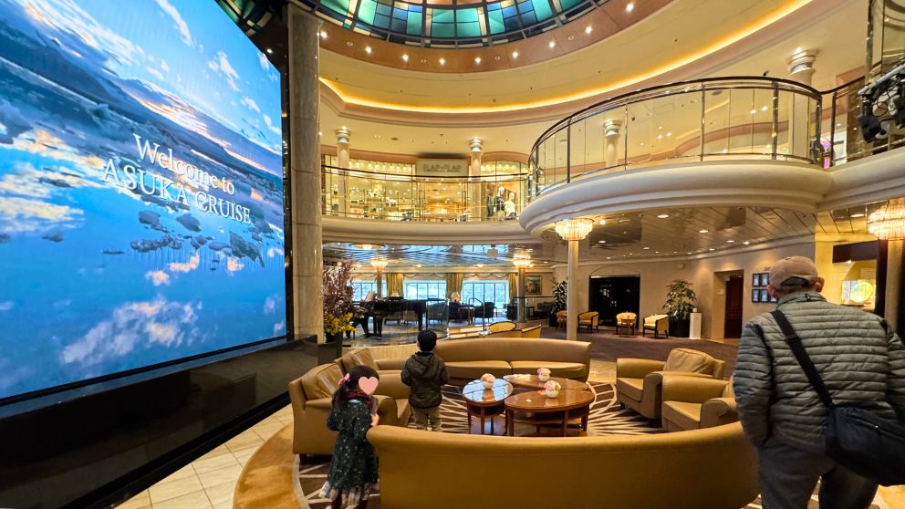 飛鳥Ⅱの豪華な吹き抜けアトリウム。天井のステンドグラスから光が差し込み、巨大LEDスクリーンには「Welcome to ASUKA CRUISE」の文字と美しい海の映像が映し出されている