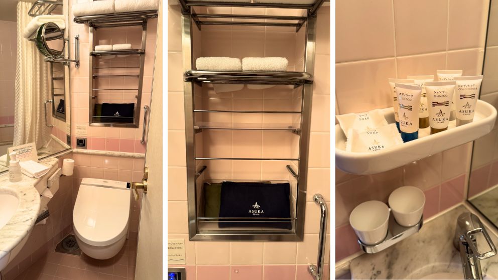 飛鳥Ⅱの客室バスルーム。ピンクのタイルと白い大理石のカウンター、洗浄機能付きトイレ、整頓されたタオル棚が見える。右側には「ASUKA CRUISE」のロゴが入ったシャンプー、コンディショナー、ボディソープ、石鹸のアメニティが並んでいる