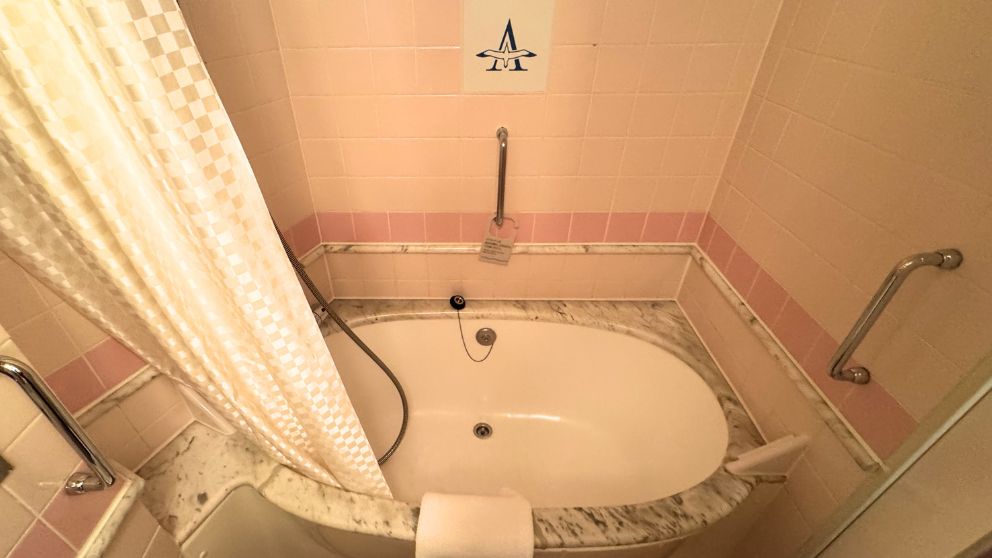 飛鳥Ⅱの客室バスルームにある、ゆったりとした白いバスタブ。壁はピンクのタイルで、天井近くには飛鳥Ⅱのアルバトロスマークが飾られている。シャワーカーテンと手すり、バスマットがセットされている