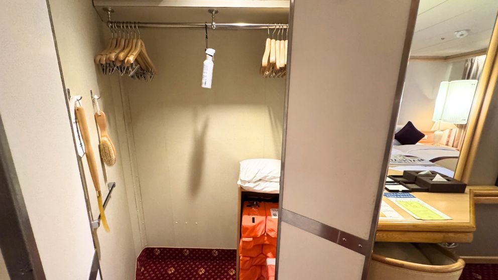 飛鳥Ⅱ客室のウォークインクローゼット内部。多数のハンガー、消臭スプレー、靴べら、衣類ブラシが掛けられている。下部の棚には、オレンジ色の救命胴衣が人数分(2着)収納されている。右側の鏡には寝室の一部が映っている