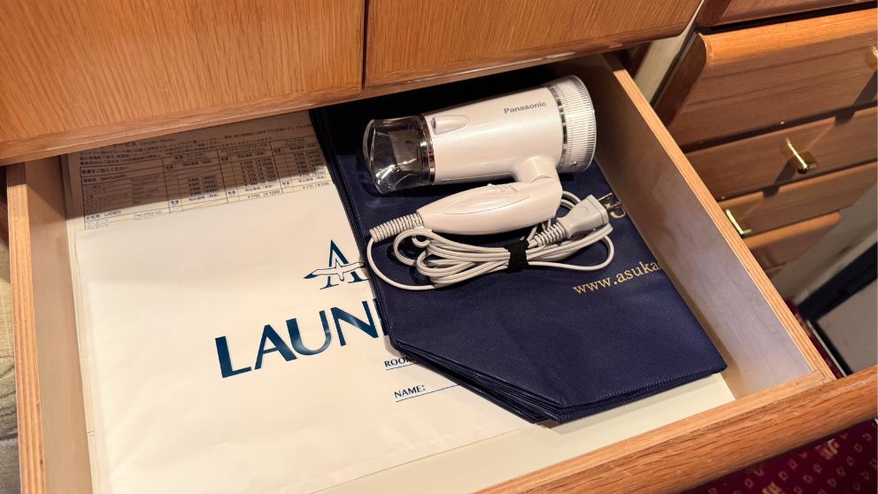 飛鳥Ⅱ客室の木製引き出し。中にパナソニック製の白いドライヤーと、飛鳥Ⅱのロゴが入ったネイビーのランドリーバッグが収納されている。