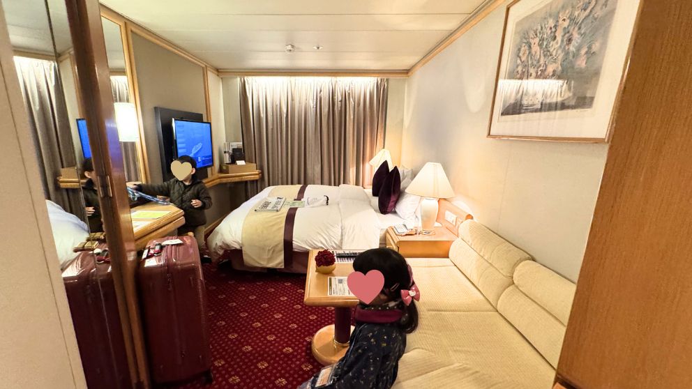 飛鳥Ⅱ客室(Fステート)の全景。赤いカーペットの部屋に、ハリウッドツインのベッド、手前に白いソファ、テレビ、デスクがある。2人の子供が部屋に入って探索しており、スーツケースが置かれている。窓にはカーテンが引かれている