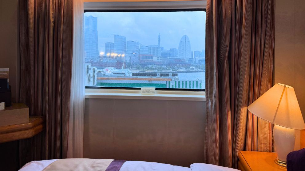 飛鳥Ⅱの客室窓から眺める、雨に濡れた横浜みなとみらいの景色。赤レンガ倉庫や大観覧車、ランドマークタワーが見える