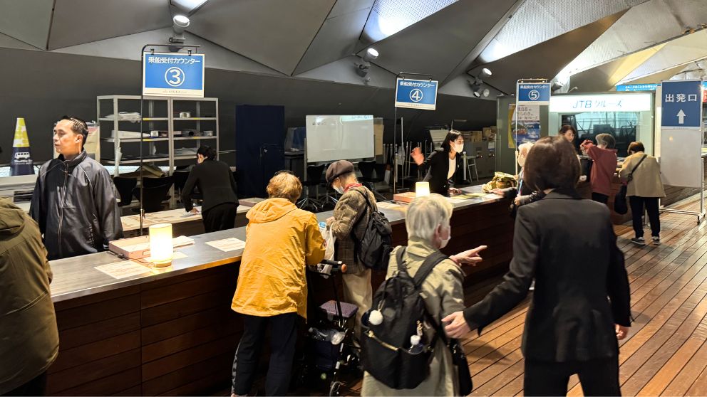 横浜港の大さん橋国際客船ターミナルにある、飛鳥Ⅱの乗船受付カウンター。「乗船受付カウンター3、4、5」の青い看板がかかり、多くの乗客が列に並んで手続きをしている。スタッフが笑顔で対応している