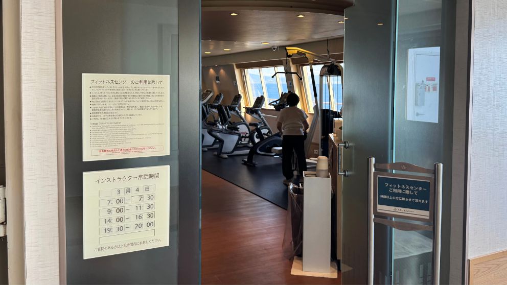 飛鳥Ⅱのフィットネスセンター入口。ガラス扉の向こうには、大海原を眺めながらトレーニングできる最新のマシンが並び、利用案内が掲示されている