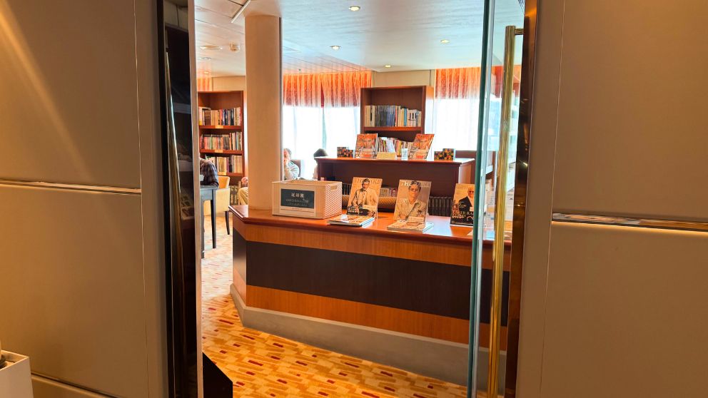 飛鳥Ⅱのライブラリーの入り口。木目調のカウンターに雑誌が並び、奥の本棚で読書をする人の姿が見える