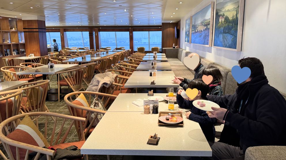カジュアルな雰囲気のリドカフェで、家族みんなで出航直後のカフェタイムを楽しむ様子。窓の外には海が見え、テーブルには思い思いに取ってきたスイーツが並ぶ