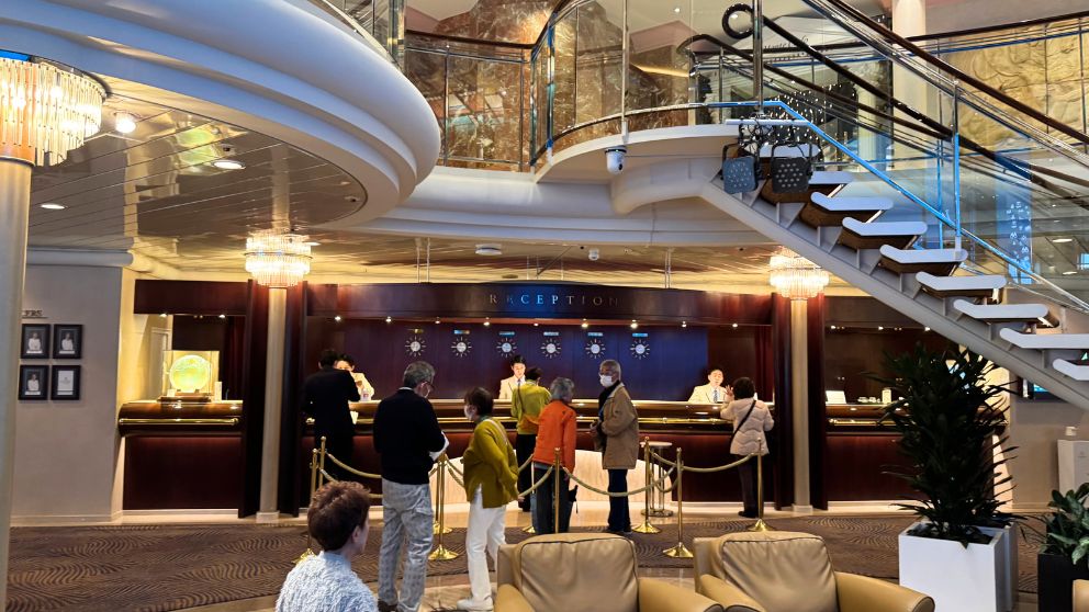 飛鳥Ⅱの豪華なレセプション(フロント)とロビー。シャンデリアが輝き、らせん階段がある吹き抜けの空間で、乗客が手続きをしている。