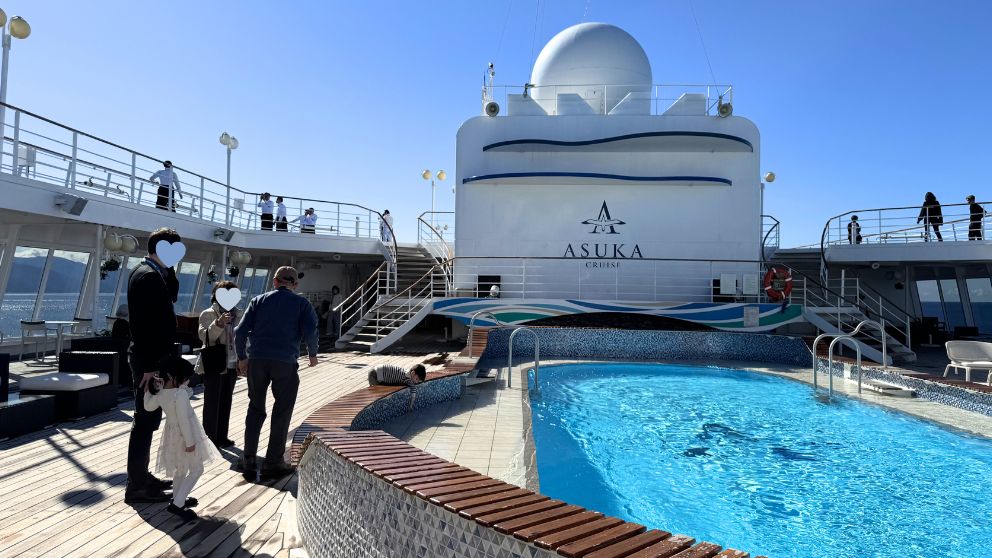 飛鳥Ⅱの11デッキ(最上階)にあるスカイプール。青空の下、乗客がプールサイドを散歩したり、景色を楽しんだりしている。ASUKA CRUISEのロゴが入った船体が美しい