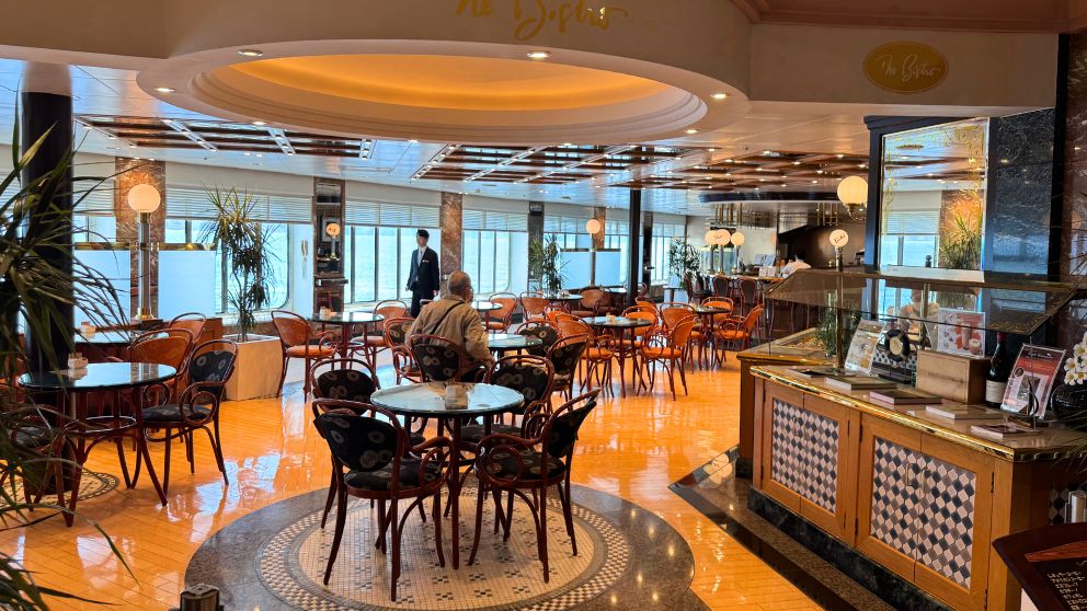 飛鳥Ⅱのラウンジ「ザ・ビストロ」の内観。オレンジを基調とした落ち着いた照明の下、エレガントな木製の椅子とテーブルが並ぶ。窓からは青い海が見え、優雅なティータイムを予感させる。