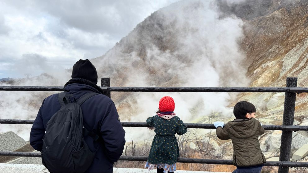 大涌谷の噴煙が上がる火山地帯を柵越しに眺める親子と、広大な谷の景色