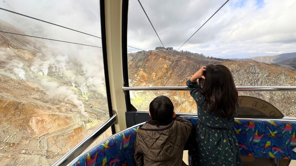 箱根ロープウェイの車内から、色づく山々の景色を夢中で眺める二人の子供の後ろ姿