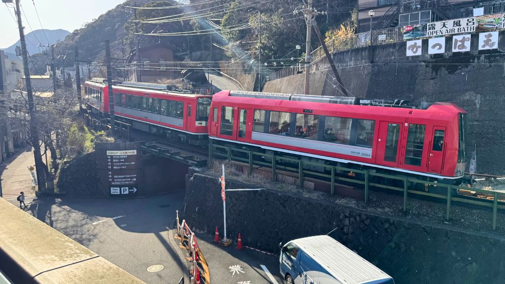 緑豊かな箱根湯本の温泉街を背景に、早川に架かる鉄橋を静かに渡る箱根登山鉄道の最新車両「アレグラ号」(赤い電車)