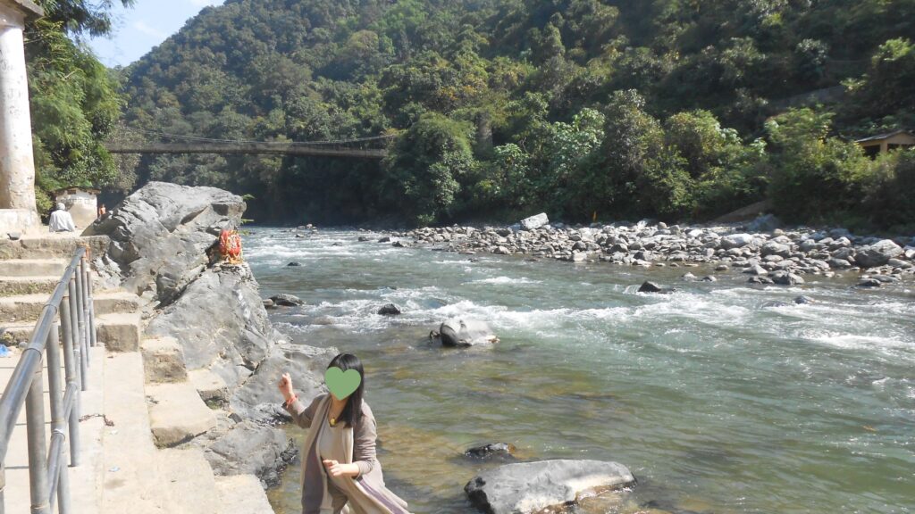 岩場のガートにいる私と、勢いよく流れるガンジス川上流、岩に祀られた小さな祠と奥に見える吊り橋
