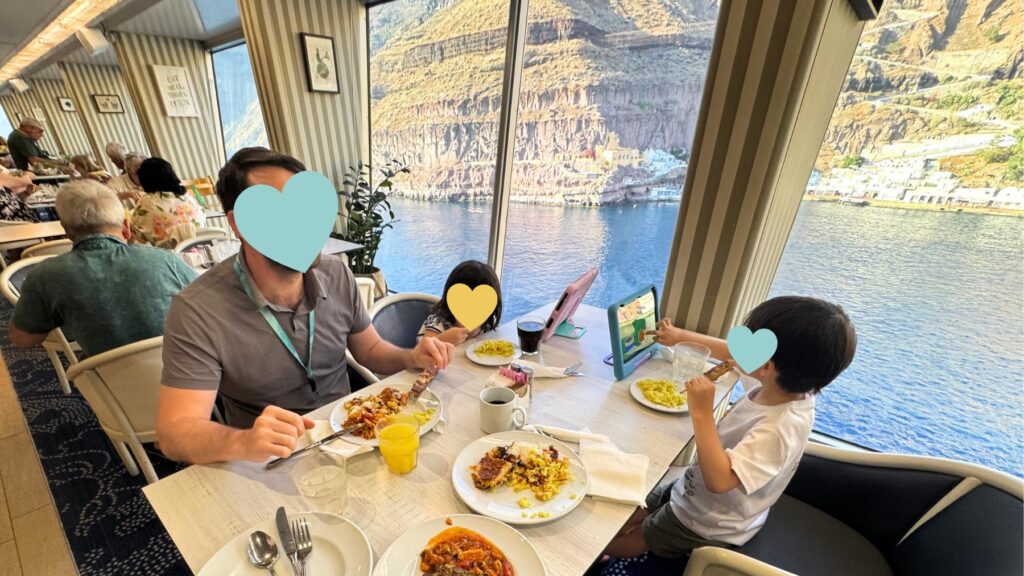 サントリーニのカルデラと崖の街並みを大きな窓から望む船内レストランで食事をとる家族