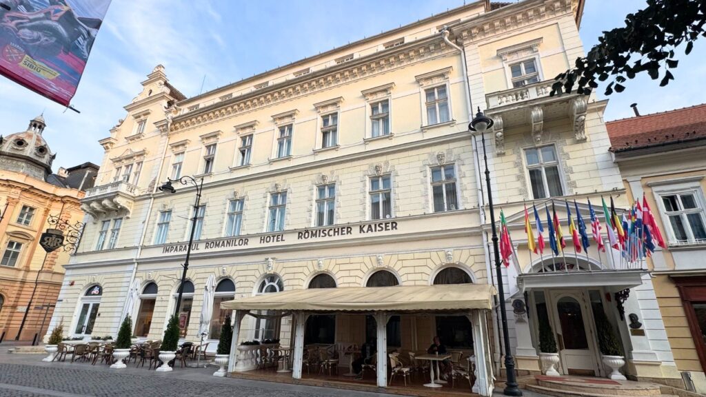 Hotel Împăratul Romanilor の外観。大広場近くに建つクリーム色のクラシックな建物で、正面に「ÎMPĂRATUL ROMANILOR HOTEL / RÖMISCHER KAISER」の文字と各国の国旗、テラス席