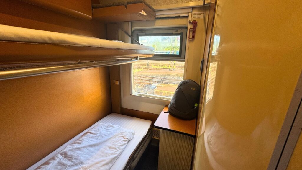 夜行列車の寝台個室。折りたたまれた寝台と窓、窓辺にリュックサックが置かれている
