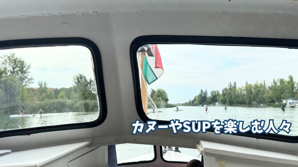 船の窓越しに見えるカヌーやSUPを楽しむ人々とハンガリー国旗