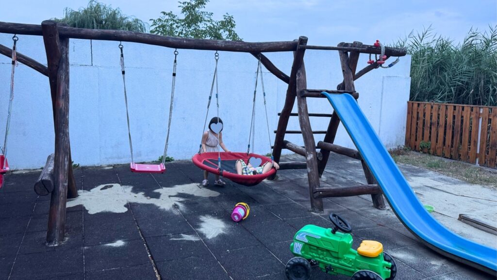 宿の遊び場で子どもをバスケットブランコに乗せる母親、青い滑り台やおもちゃのトラクター