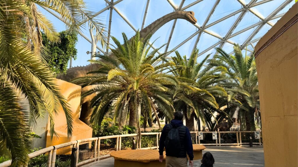 Biodóm内の植物の上に長い首を伸ばす恐竜模型と通路を歩く来園者
