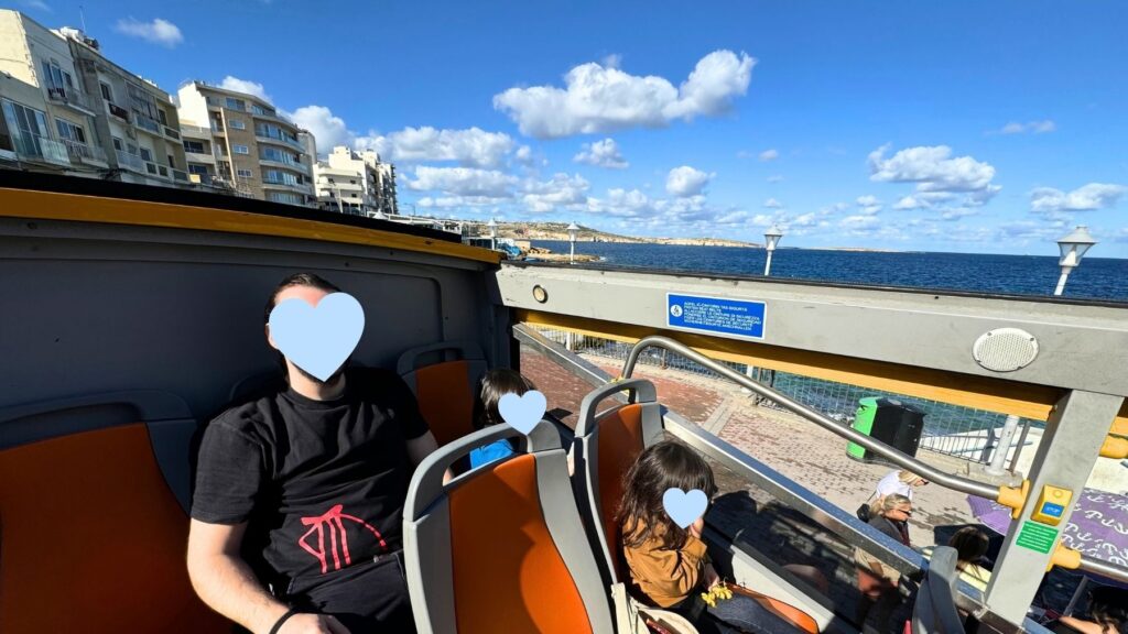 ホップオン・ホップオフバスの2階席に座る家族。車窓には海沿いの町並みと青い海が広がる。